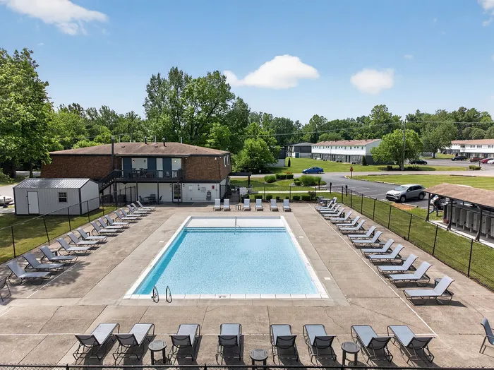 Inviting community pool area with ample lounge chairs, surrounded by lush greenery and cozy residential buildings.