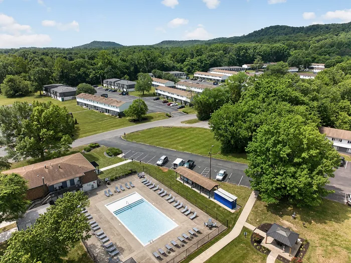 Vibrant aerial view of a scenic apartment complex with lush greenery, a sparkling swimming pool, and spacious parking areas.