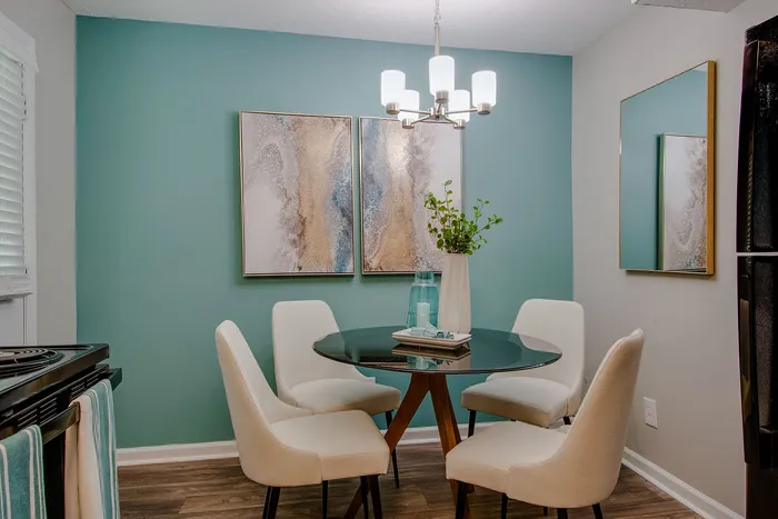 Chic dining nook featuring a stylish glass table and modern chairs against a calming teal accent wall, complemented by elegant artwork and soft lighting.