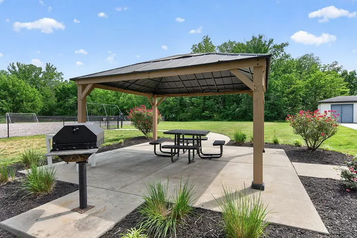 Charming outdoor gazebo with picnic table and BBQ grill, surrounded by lush greenery and vibrant flowers, ideal for outdoor relaxation.