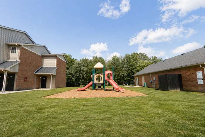 Charming community playground with vibrant red slides, surrounded by lush greenery and apartments under a clear blue sky.