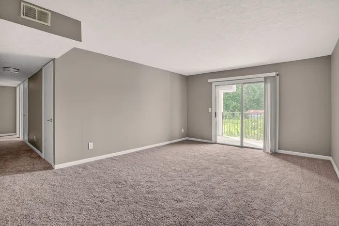 Bright and spacious living room with plush carpet and sliding glass doors leading to a serene balcony view.
