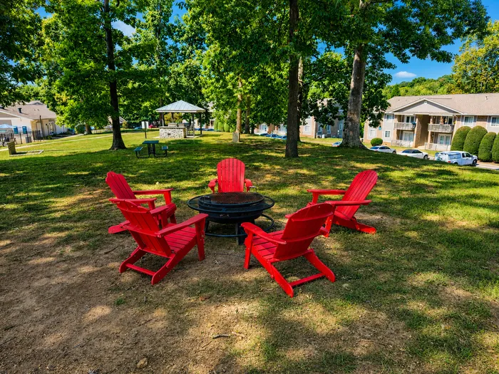 Charming outdoor area with vibrant red Adirondack chairs around a cozy fire pit, surrounded by lush greenery and nearby apartment buildings.