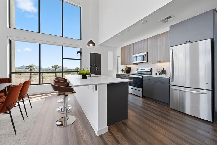 Callia Kitchen, Large windows, High ceiling, wood flooring, stainless steel appliances, light fixture, kitchen isalnd with barstools, wooden cabinets