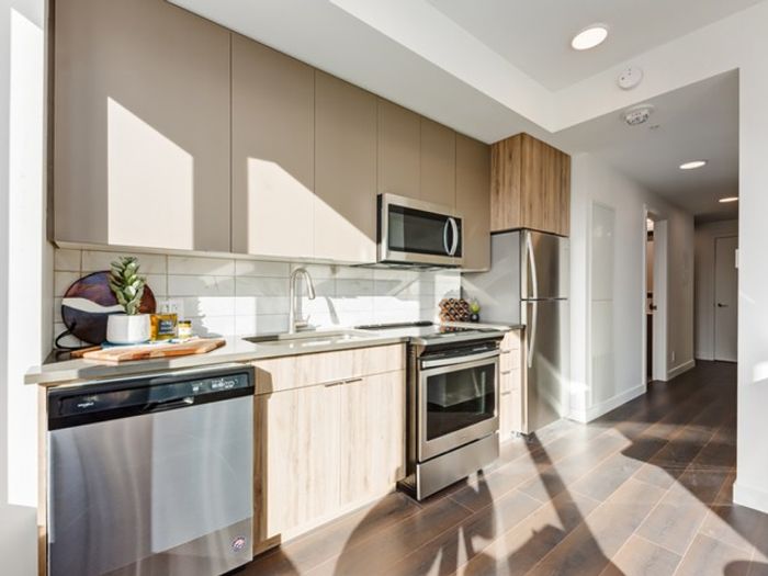 A bright, modern kitchen at One Park featuring stainless steel appliances, light wood cabinetry, white backsplash, and stone countertops.