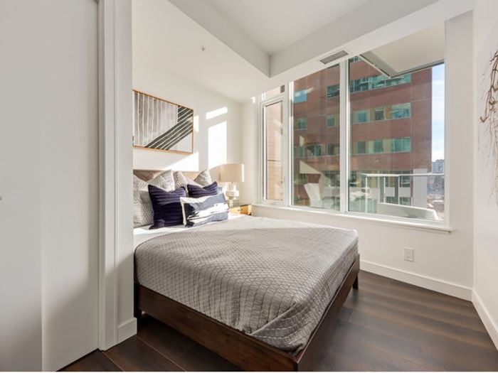 A modern One Park bedroom featuring a large window with city views, a cozy bed, hardwood floors, and neutral-toned decor.