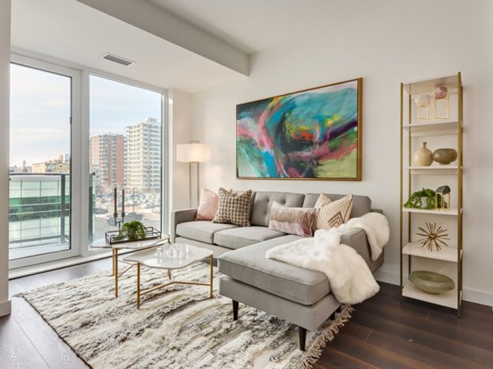 A cozy, sunlit living room at One Park featuring a patterned area rug, modern sofa, glass coffee table, tall bookshelf, and sliding doors opening to a private outdoor space.