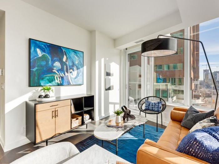 Modern apartment living room with a tan leather sofa, desk setup, wood sideboard, large wall art, and full-height windows overlooking the city.