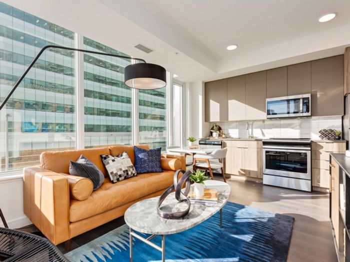 Contemporary living room with a tan leather sofa, round coffee table, blue accent rug, wall-mounted TV, and large windows bringing in natural light.