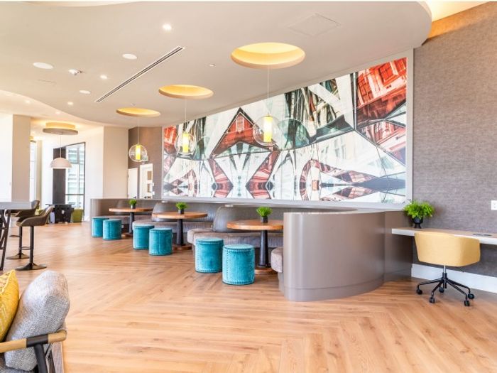 Modern co-working lounge with abstract mural, circular tables, teal stools, and a built-in desk along the wall beneath warm pendant lights.