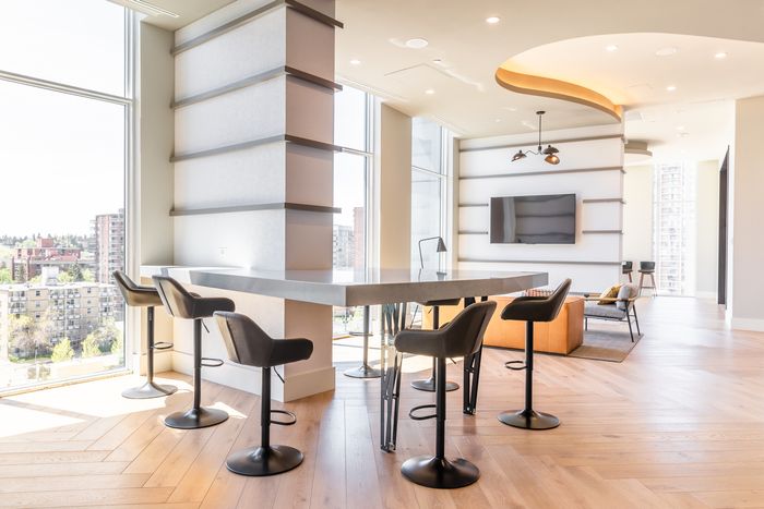 Modern high-top tables and barstool seating arranged near full-length windows in a bright coworking lounge with wood flooring and integrated shelving.