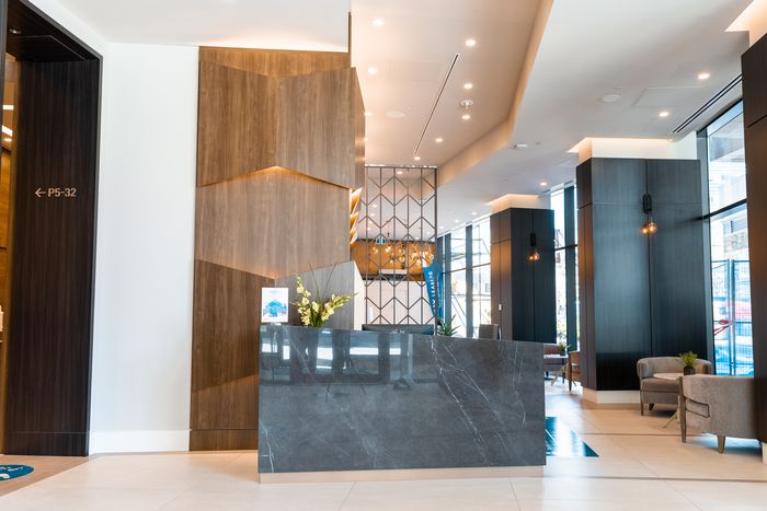 Contemporary apartment lobby featuring a marble reception desk, geometric wood accent wall, and cozy seating area with floor-to-ceiling windows.