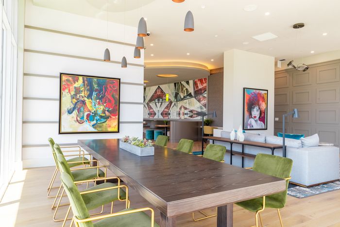 A stylish lounge space featuring a long wood table with green velvet chairs, vibrant artwork, and modern lighting fixtures.