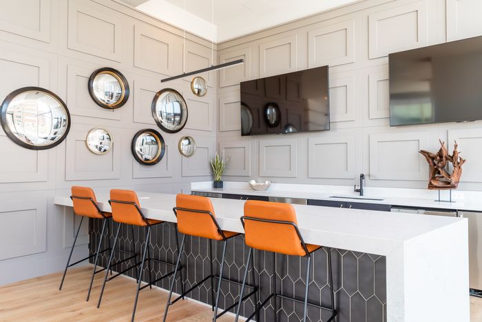 Modern community kitchen with bar seating, wall-mounted mirrors, and sleek finishes, featuring leather barstools and a mounted TV.