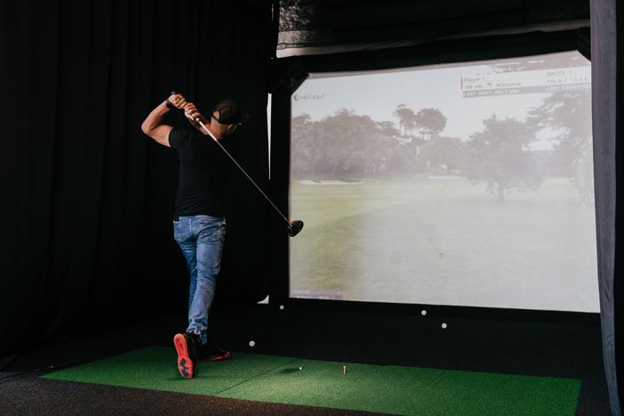 Person swinging a golf club in front of a large indoor simulator screen displaying a digital golf course.