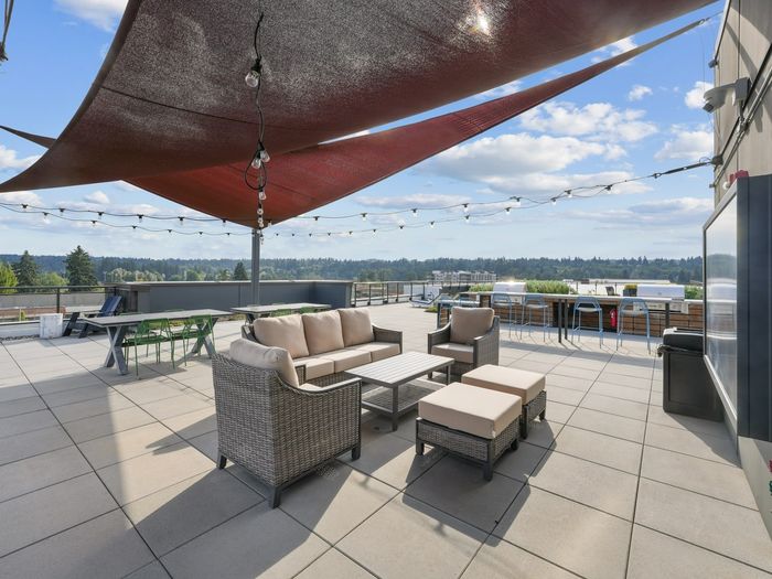 Rooftop terrace featuring cushioned wicker seating, dining tables, bar seating, and overhead string lights beneath shade sails.