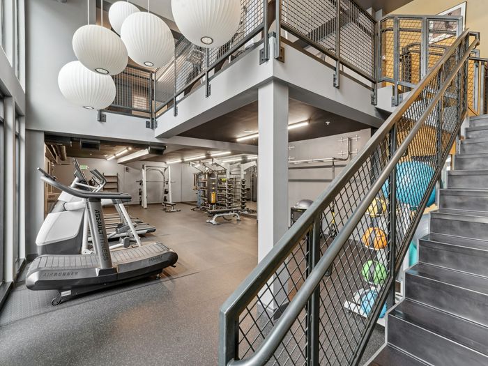 Spacious two-level apartment gym with treadmills, strength machines, free weights, medicine balls, and a stairway leading to an upper workout area.