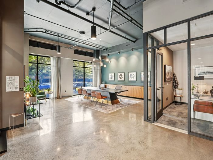 Stylish lounge with a large conference table, leather chairs, modern light fixtures, floor-to-ceiling windows, and a private glass-enclosed office