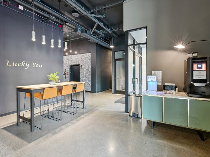 Contemporary apartment lobby featuring a black accent wall with &ldquo;Lucky You&rdquo; text, high-top table with orange barstools, modern pendant lighting, and a coffee and water station near a glass-walled office.