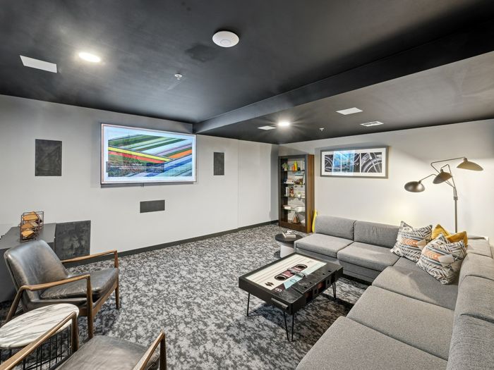 Stylish media room with a large sectional sofa, modern accent chairs, a mounted flat-screen TV, bookshelf, floor lamp, and decorative art on the walls.