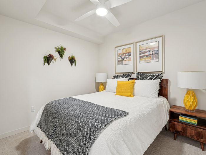 Cozy bedroom featuring a white bedspread with black and yellow accent pillows, framed abstract artwork, yellow lamps, wood nightstands, and hanging wall planters