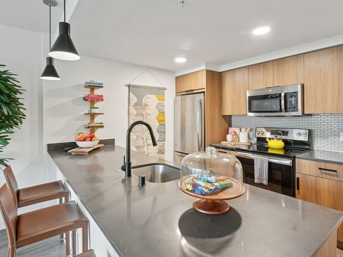Stylish kitchen featuring wood cabinetry, stainless steel appliances, a gray quartz island with black fixtures, barstools, pendant lighting, and decorative wall art