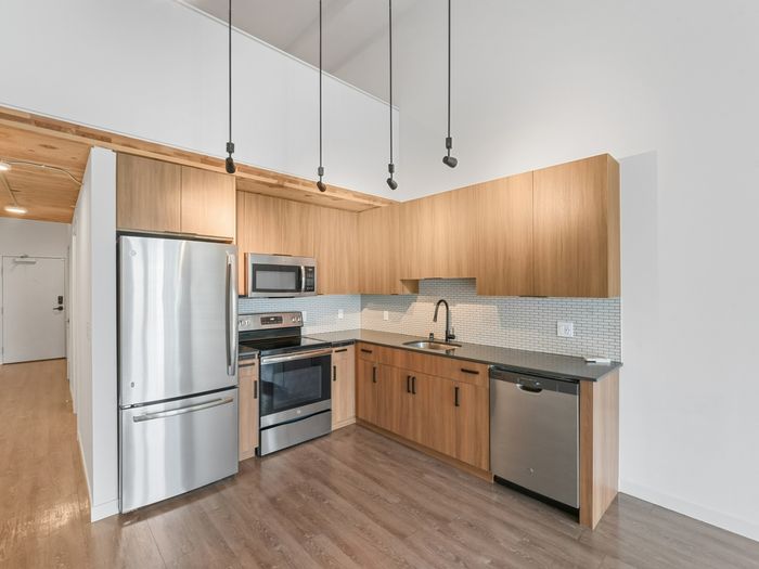 Modern apartment kitchen with wood cabinetry, stainless steel appliances, quartz countertops, a tiled backsplash, and hanging pendant lights