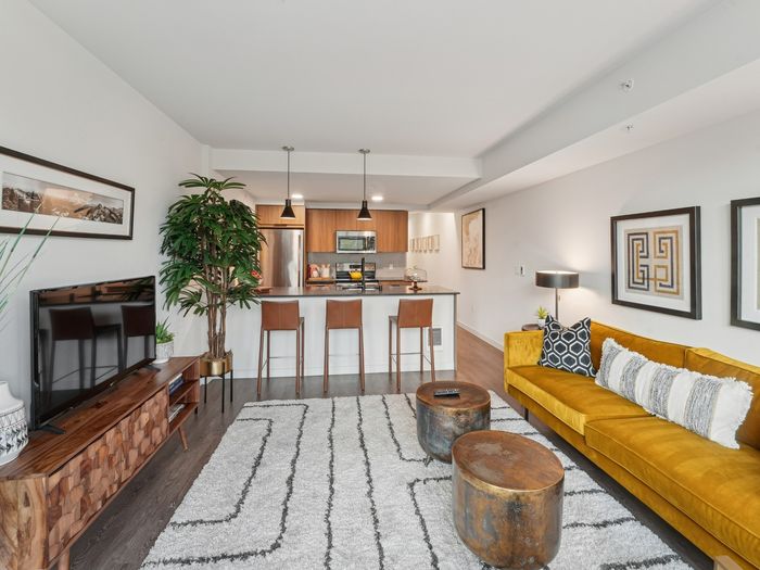 Contemporary living space featuring a mustard yellow sofa, TV console, barstools at a kitchen island, pendant lighting, and wood cabinetry in the background.