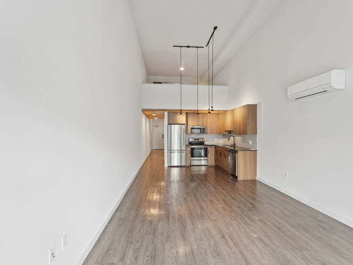 Unfurnished apartment with tall white walls, wood-look flooring, stainless steel appliances, wood cabinetry, hanging light fixtures, and a wall-mounted air conditioning unit.