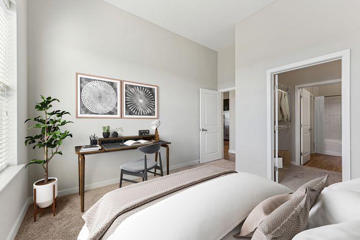 Modern bedroom with a neatly made bed, a wooden desk and chair by the window, indoor plant, two framed artworks on the wall, and an open walk-in closet leading to a bathroom.