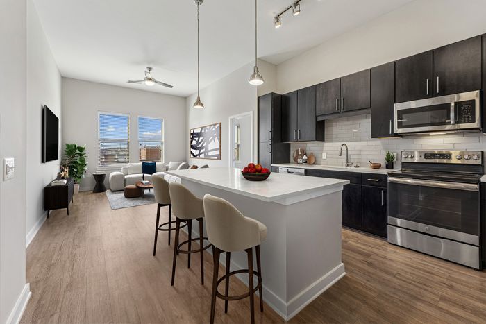 Modern kitchen with dark cabinets, stainless steel appliances, and a large white island with bar stools, opening into a bright living room with wood flooring and two windows.