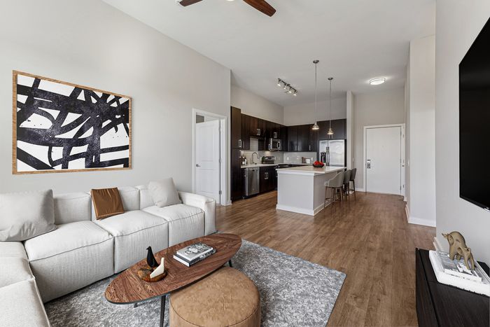Contemporary open-concept living room and kitchen with wood floors, a white sectional sofa, dark cabinets, a large island with pendant lights, and abstract wall art.