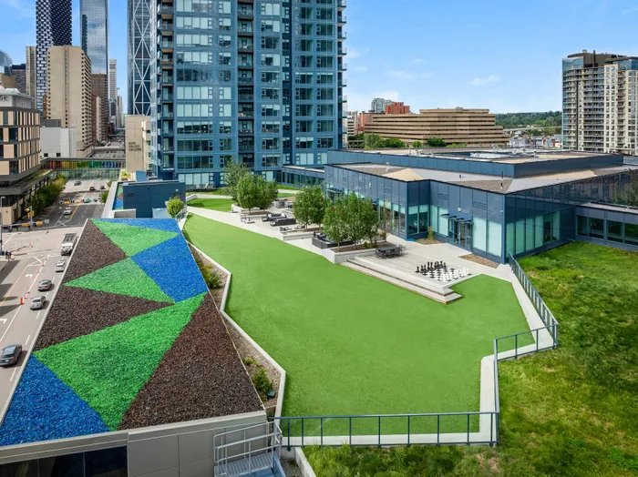 Vibrant urban rooftop terrace with colorful geometric designs, featuring a lush green lawn and outdoor seating area, surrounded by modern high-rise buildings.