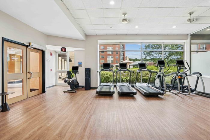 Bright and modern fitness center with large windows, featuring treadmills and exercise bikes on polished wooden floors, offering a vibrant workout environment.