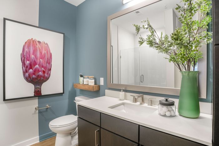 Chic bathroom with elegant blue walls, modern fixtures, and vibrant art decor, featuring a large mirror and stylish green vase with lush greenery.