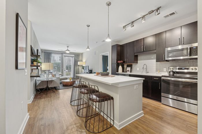 Chic open-concept living space featuring a modern kitchen with dark cabinetry, sleek appliances, and a spacious breakfast bar leading into a cozy living room with elegant decor and natural light.