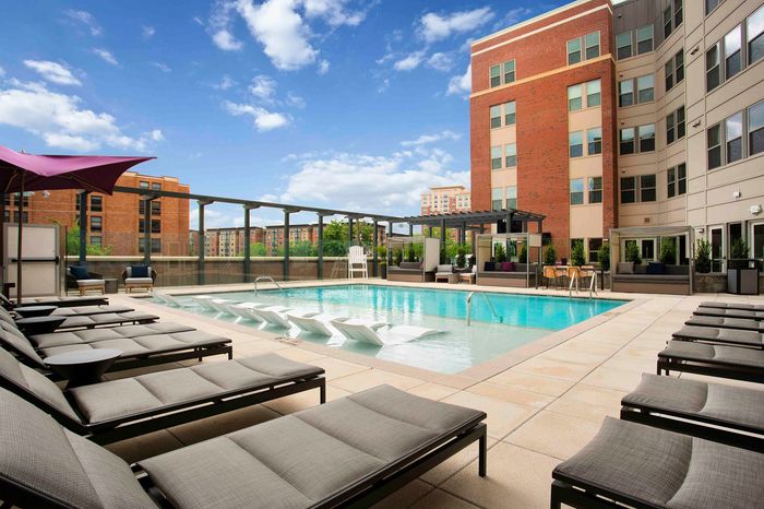 Luxurious apartment complex featuring a sparkling outdoor pool with modern lounge chairs, shaded seating areas, and vibrant city views, perfect for relaxation and leisure.