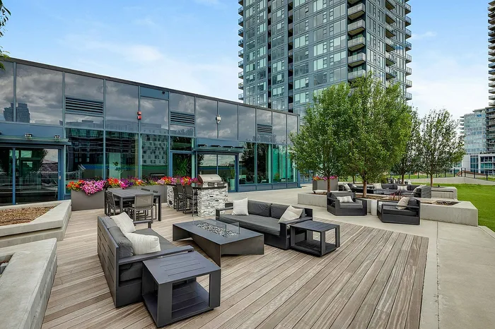 Modern outdoor terrace with stylish seating, a barbecue area, and vibrant flower arrangements, set against a striking glass building and lush greenery.