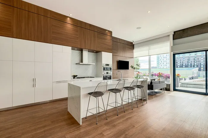 Modern kitchen, dining, and living area with sleek wood cabinetry, marble island, stylish bar stools, and large windows overlooking a vibrant balcony garden.