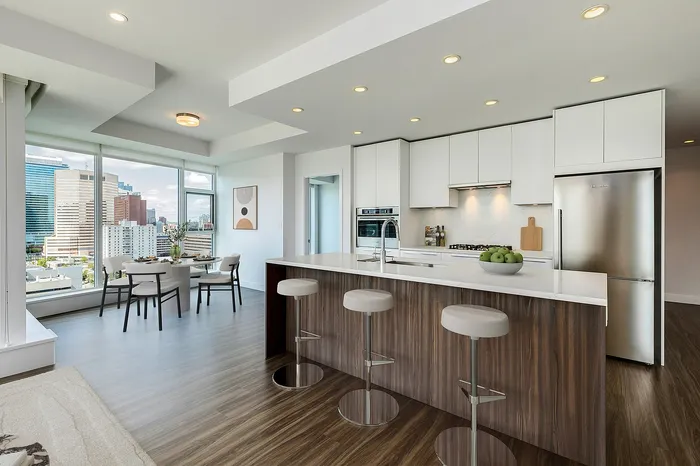 Modern apartment interior featuring a sleek kitchen island with bar stools, open-concept dining area, and stunning city views through large windows.