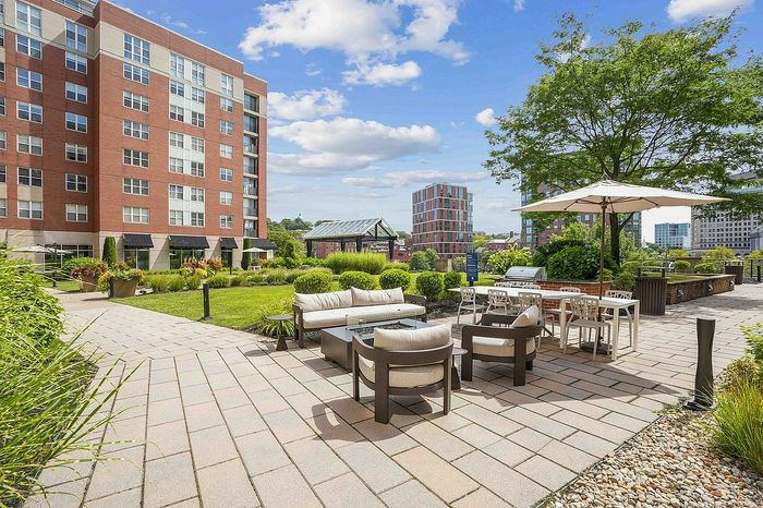 Vibrant courtyard with modern outdoor seating, lush greenery, and a charming brick apartment building backdrop under a clear blue sky.