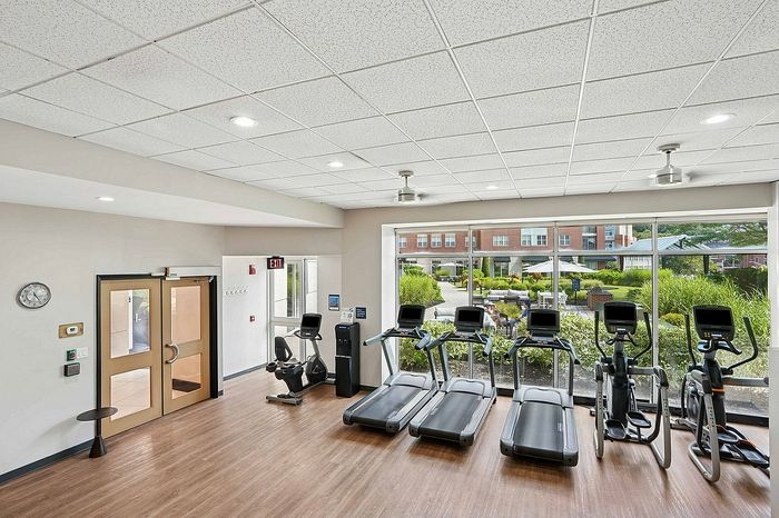 Modern fitness room with large windows, featuring treadmills and exercise bikes, overlooking beautifully landscaped gardens.