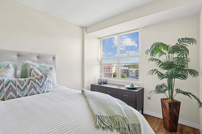Bright and serene bedroom featuring a plush bed with stylish pillows, large window with a cityscape view, cozy throw blanket, and elegant indoor plant.