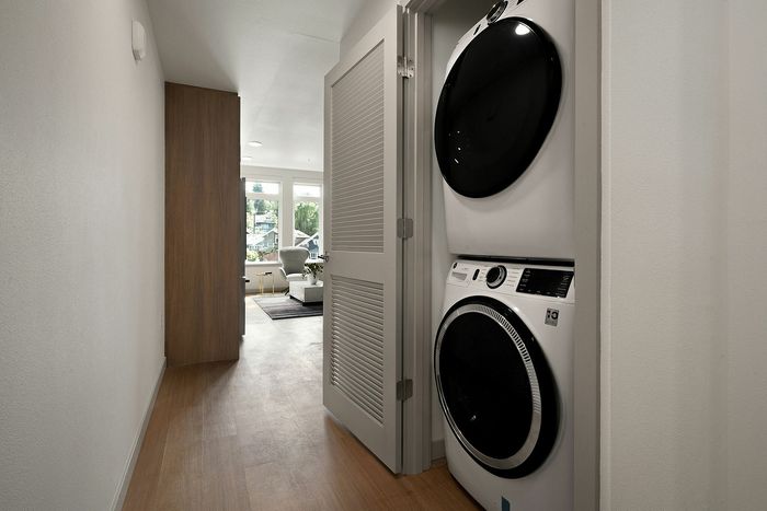 Modern apartment hallway with sleek stacked washer and dryer, leading to a bright, inviting living area with large windows overlooking greenery.