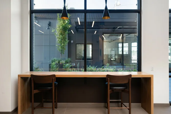 Modern workspace with a sleek wooden desk and chairs, overlooking lush greenery through large windows, complemented by stylish pendant lighting.