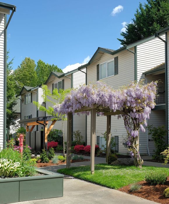 Charming apartment complex exterior featuring flowering pergola, vibrant landscaping, and bright, sunny facade.