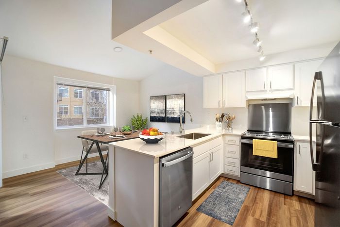 Modern apartment kitchen with sleek white cabinets, stainless steel appliances, and a bright dining area by a large window. Perfect for entertaining and everyday living.