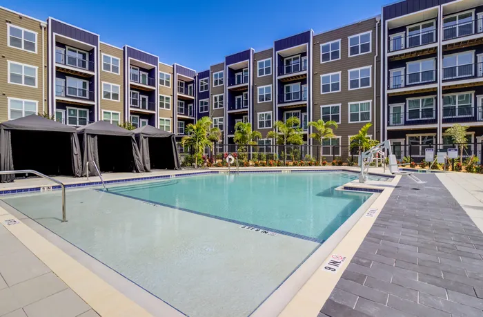 A bright photo showing large pool with crystal blue water, a sundeck, and cabanas, with young palm trees and a new apartment complex in the background.