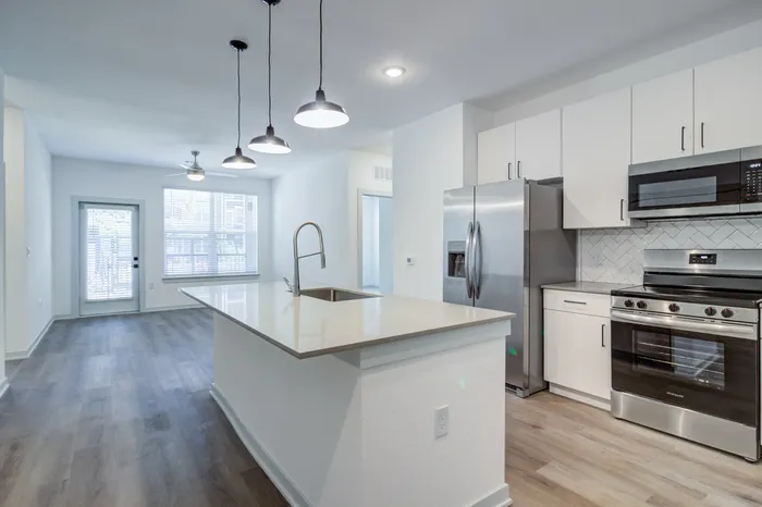 Unfurnished open-concept apartment kitchen flowing into the living room with large windows, patio access, stainless steel appliances, white cabinets, and wood-style plank flooring at Garrett Forum in Fort Myers, Florida.