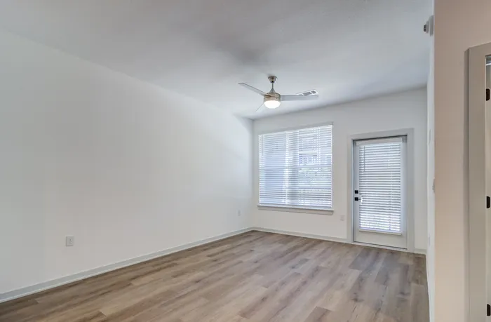 Unfurnished apartment living room with wood-style plank flooring, ceiling fan, large window, and private patio or balcony access at Garrett Forum in Fort Myers, Florida.